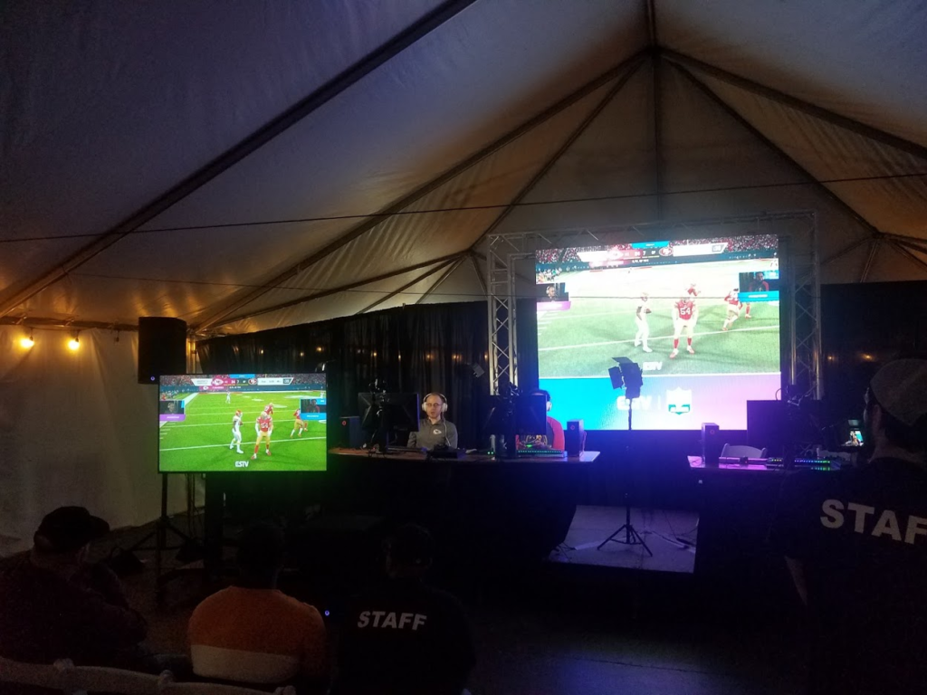 Two large screens show video game play during a gaming event inside a large tent.